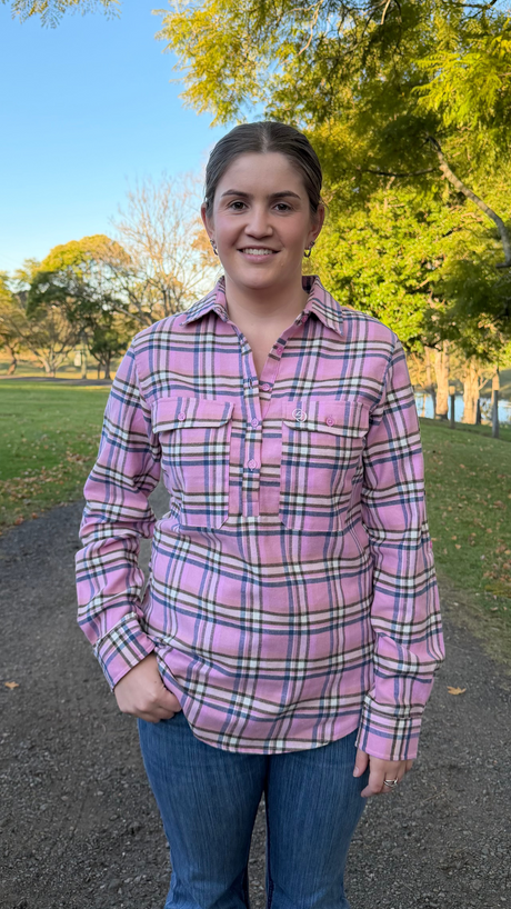 A woman standing outdoors on a gravel path wearing a pink plaid button-up shirt and blue jeans, with trees and grass in the background.