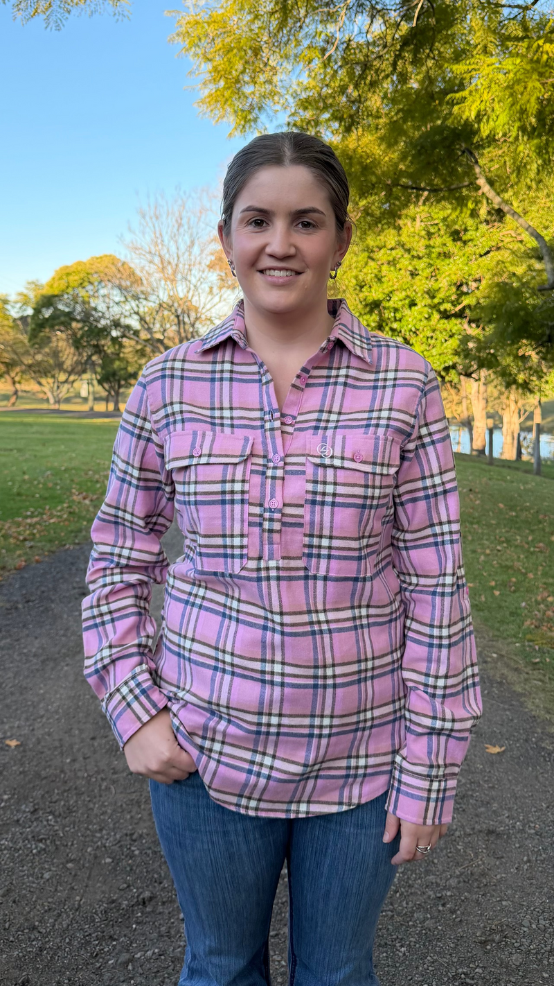 A woman standing outdoors on a gravel path wearing a pink plaid button-up shirt and blue jeans, with trees and grass in the background.