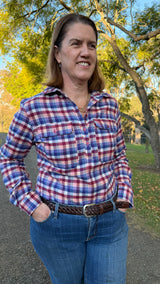A woman wearing a red, blue, and white plaid button-down shirt with chest pockets, paired with blue jeans and a brown braided belt, standing outdoors in a park with trees in the background.