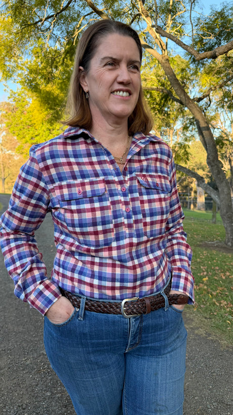 A woman wearing a red, blue, and white plaid button-down shirt with chest pockets, paired with blue jeans and a brown braided belt, standing outdoors in a park with trees in the background.
