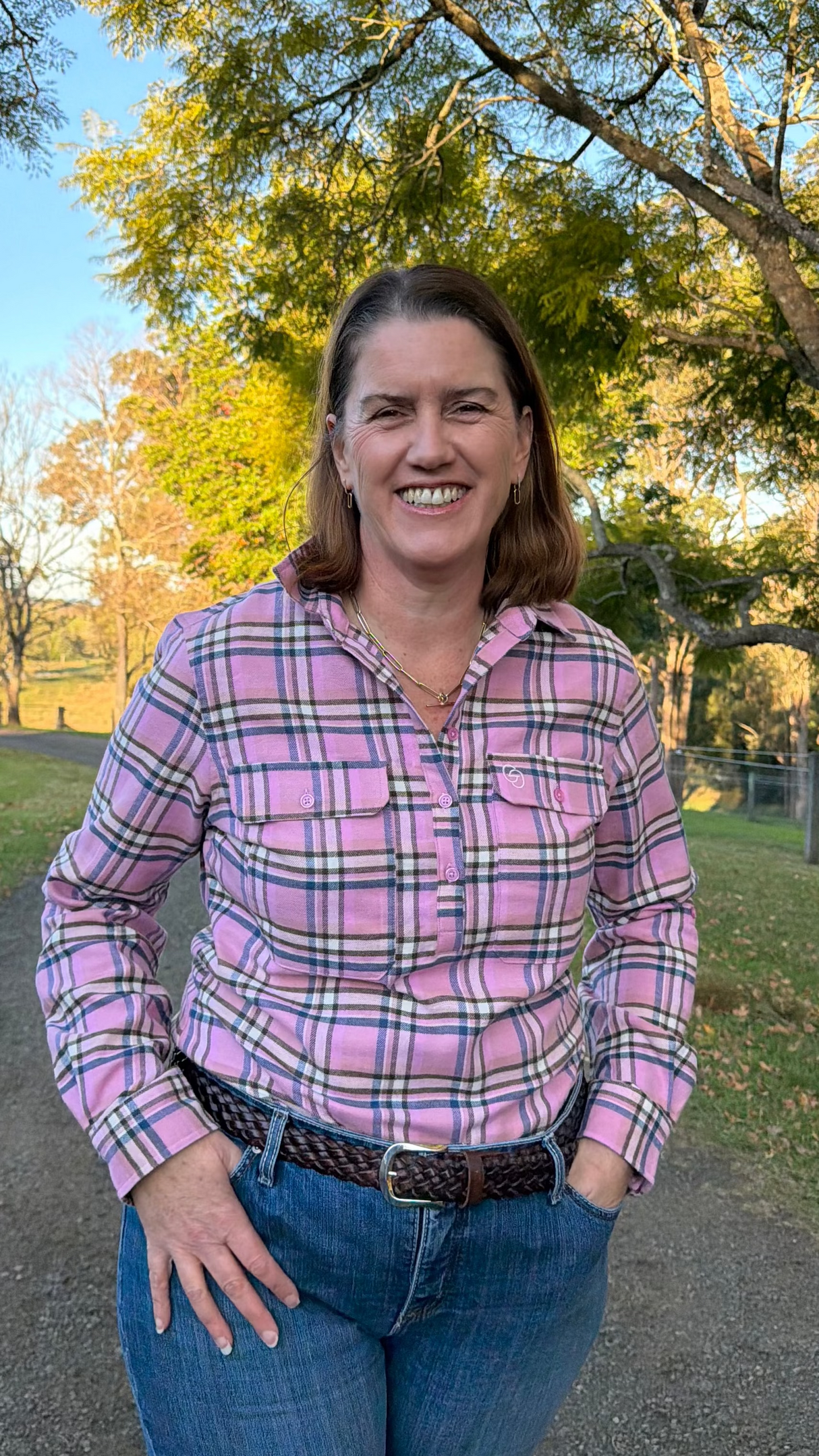 A woman smiling outdoors on a dirt path wearing a pink plaid button-down shirt and blue jeans, with trees and grass in the background.
