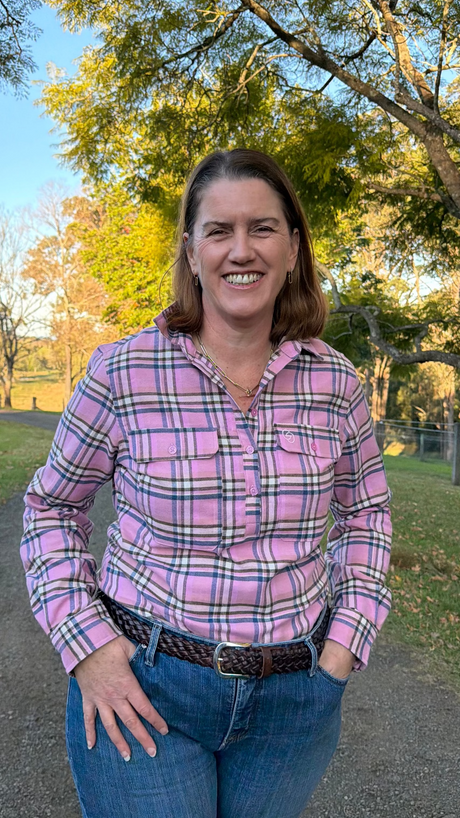 A woman smiling outdoors on a dirt path wearing a pink plaid button-down shirt and blue jeans, with trees and grass in the background.