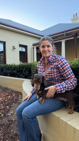 A woman sitting on a low wall in front of a house, wearing a red, blue, and white plaid flannel shirt and blue jeans, holding a small brown dachshund.