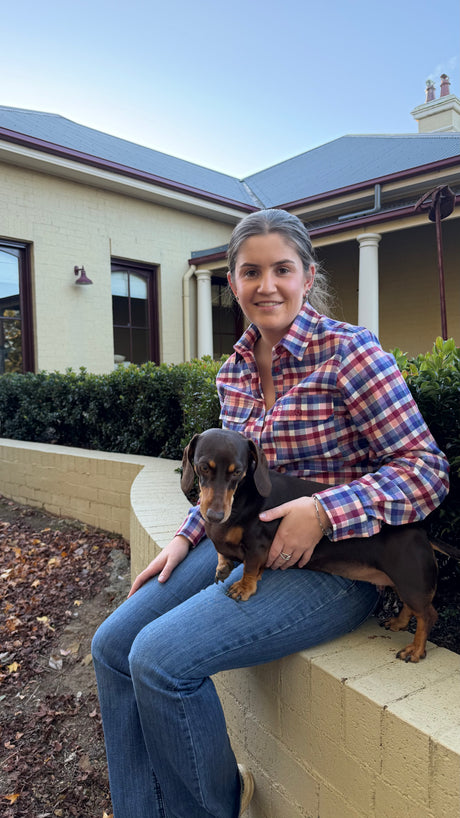 A woman sitting on a low wall in front of a house, wearing a red, blue, and white plaid flannel shirt and blue jeans, holding a small brown dachshund.