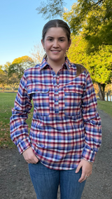 A woman standing outdoors on a dirt path wearing a red, white, and blue plaid long-sleeve button-up shirt and blue jeans, with trees and grass in the background.