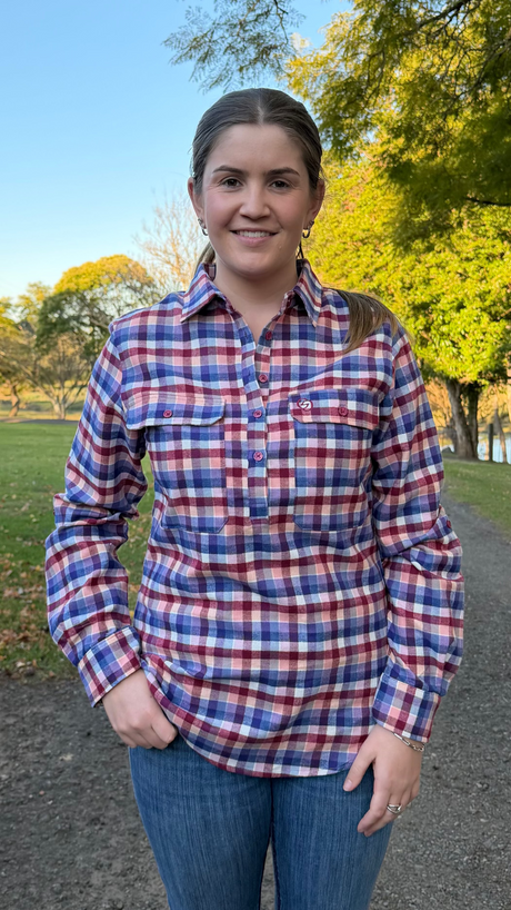A woman standing outdoors on a dirt path wearing a red, white, and blue plaid long-sleeve button-up shirt and blue jeans, with trees and grass in the background.