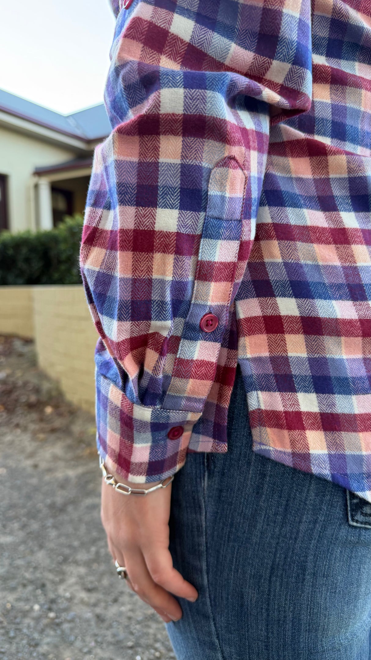 A close-up of a person wearing a long-sleeve plaid button-up shirt in red, blue, and cream, paired with blue jeans, with a blurred outdoor background.