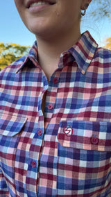 Close-up of a person wearing a plaid button-up shirt in red, blue, and white, with a small circular logo patch on the left chest pocket. The background shows a blurred outdoor scene with blue sky and greenery.