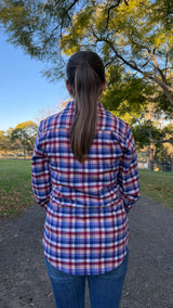 A woman viewed from behind wearing a red, blue, and white plaid button-up shirt and blue jeans, standing on a dirt path in a park with trees and blue sky in the background.