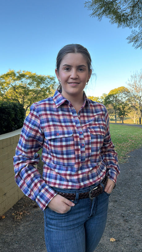 A woman wearing a red, blue, and white plaid button-down shirt and blue jeans, standing outdoors on a path with trees and grass in the background.