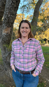 A smiling woman standing outdoors in front of trees, wearing a pink plaid button-up shirt with blue jeans and a brown belt.
