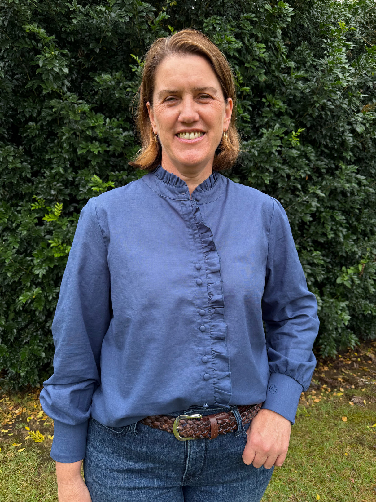A woman smiling outdoors in front of a green hedge, wearing a blue long-sleeve blouse with a gathered ruffle collar and front placket, paired with blue jeans and a brown woven belt.