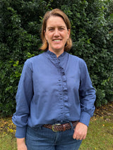 A woman smiling outdoors in front of a green hedge, wearing a blue long-sleeve blouse with a gathered ruffle collar and front placket, paired with blue jeans and a brown woven belt.