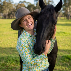 Woman in a floral shirt and wide-brimmed hat petting a horse in a field.