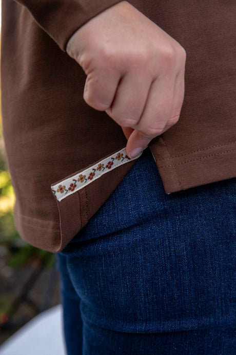 Close-up of a lady wearing a chocolate brown rugby with a floral label/tape, standing outdoors.